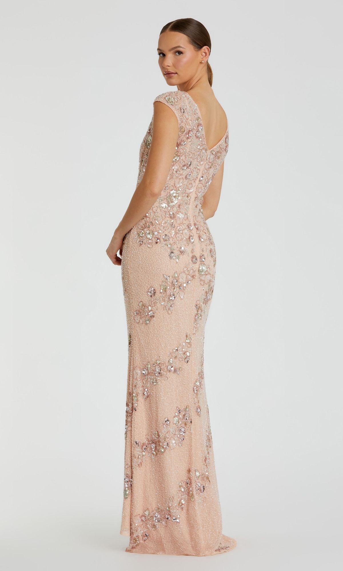 A woman poses in the Mac Duggal 5500 Beaded Long Formal Dress with cap sleeves, floor-length in pale pink, glancing over her shoulder against a plain white background.